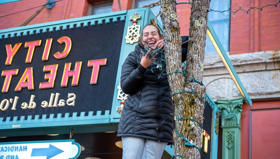 A female student wraps lights around the trunk of a tree in front of Biddeford's City Theater